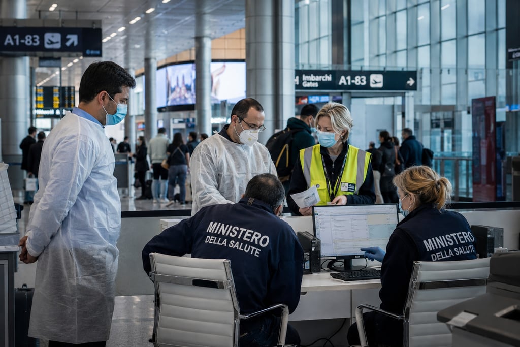 Italian health ministry staff in masks conduct passenger screenings at a busy airport terminal.