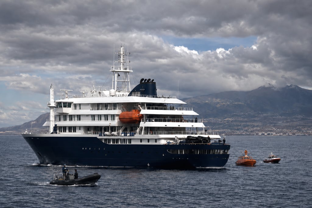A luxury cruise ship anchored in deep blue water near a mountainous coastline with small escort boats.