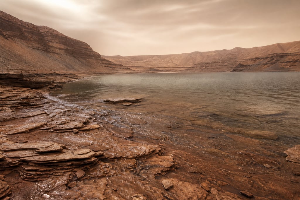 Layered rock formations lining a desert lake under a hazy sky in a canyon landscape.