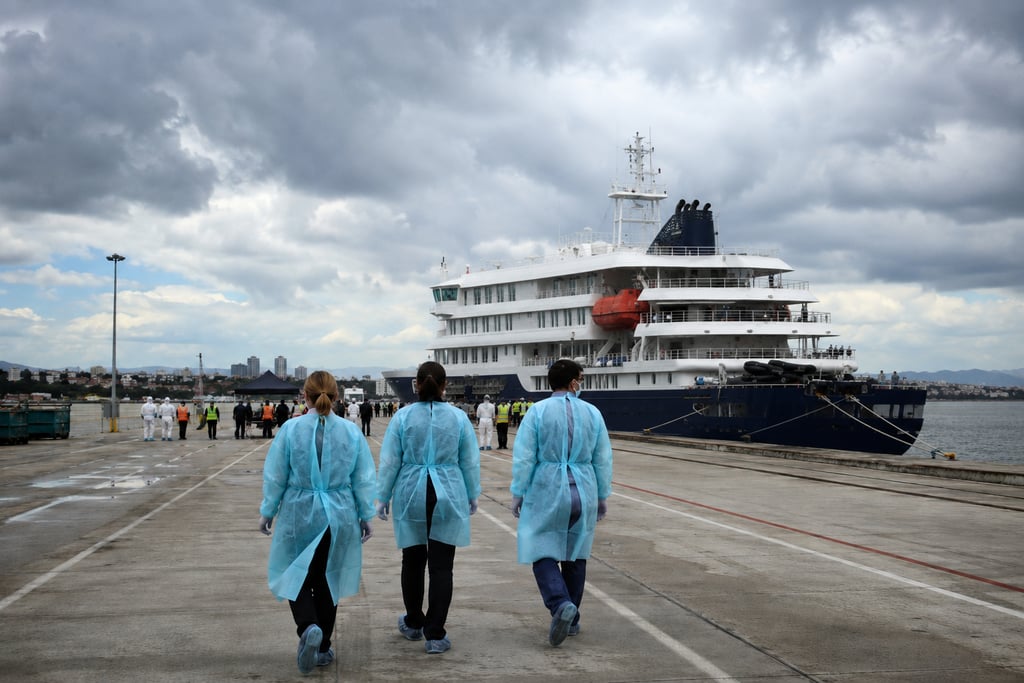 Medical workers in blue protective gowns walking toward a large cruise ship docked at a port terminal.
