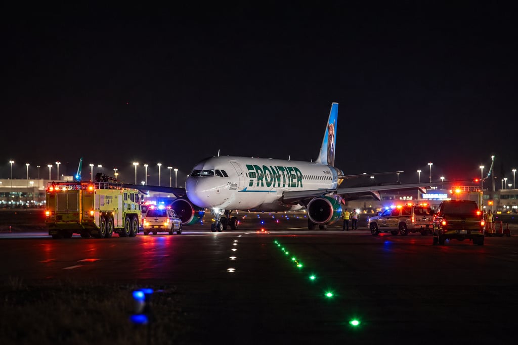 A Frontier Airlines plane on an airport runway at night surrounded by emergency fire and rescue vehicles with flashing