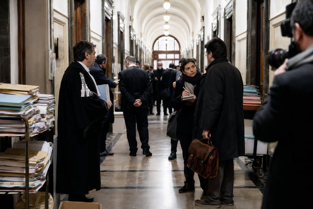 A lawyer in a black robe stands in a courthouse hallway near a journalist interviewing a man.