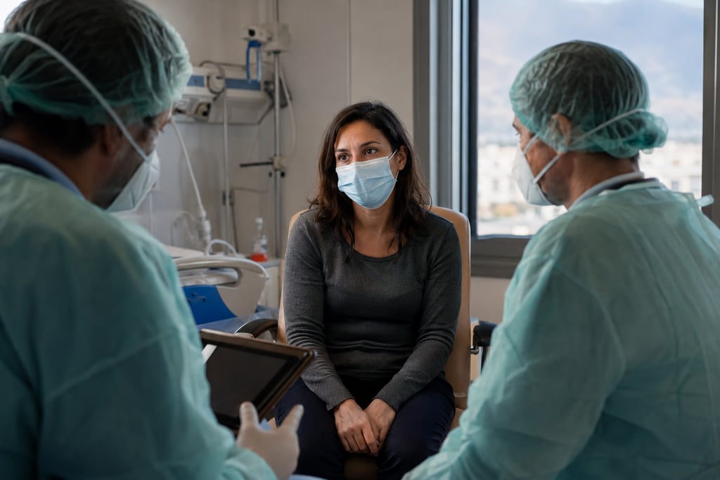 Female patient wearing a face mask discussing medical treatment with doctors in a hospital room.