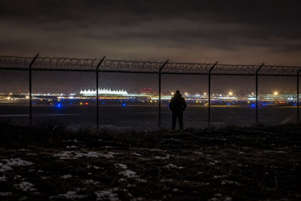 Silhouette of a person looking through a chain-link fence at a bright airport runway at night.