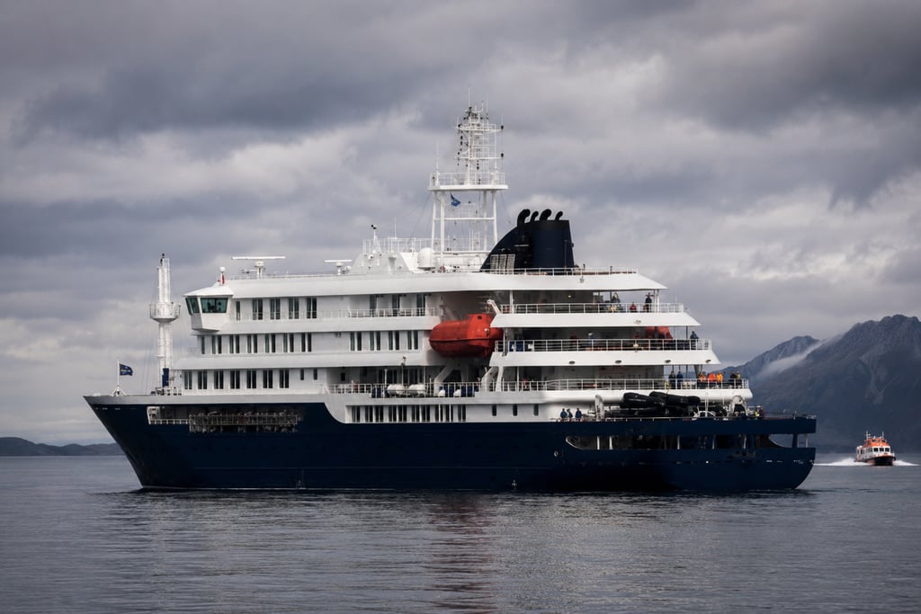 Luxury expedition cruise ship sailing on calm waters near mountains under a cloudy sky.