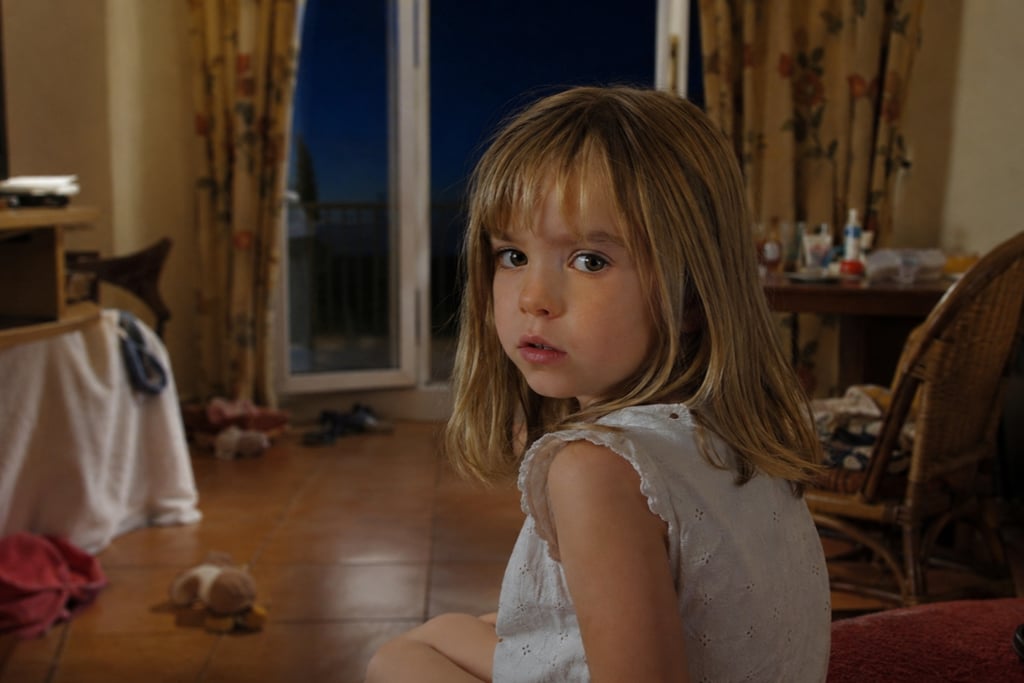 A young blonde girl looking over her shoulder inside a dimly lit room with an open balcony door.