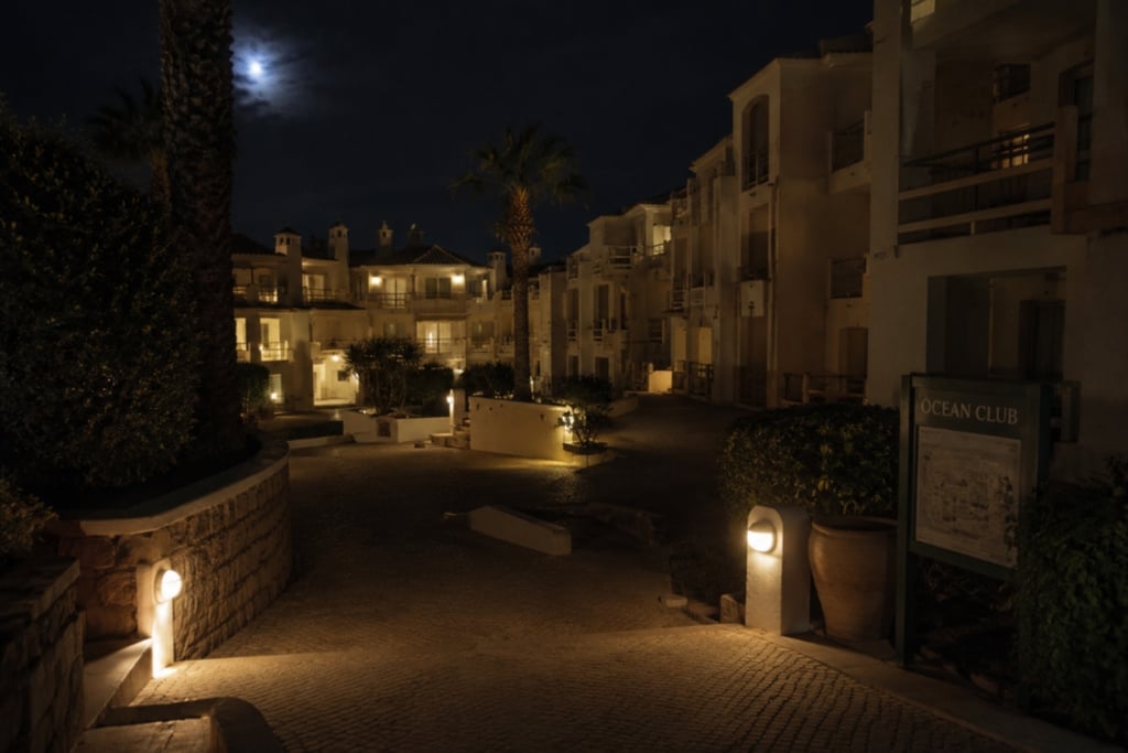 Night view of a luxury ocean club resort with lit stone pathways, white buildings, and a full moon.