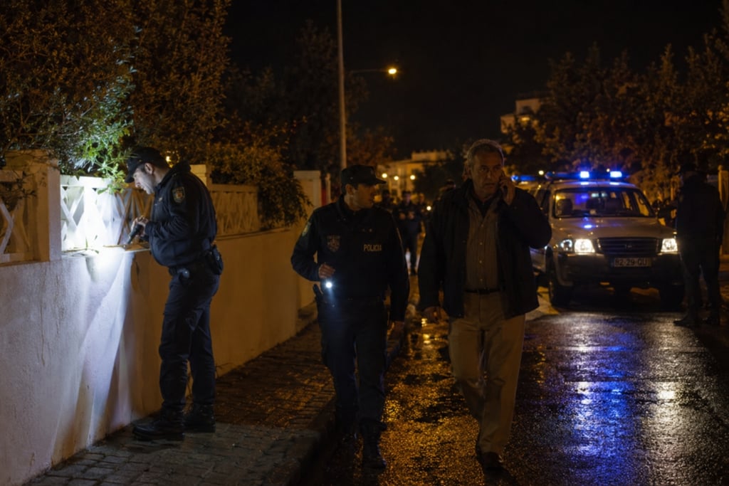Police officers investigate a crime scene at night on a wet street with patrol cars.