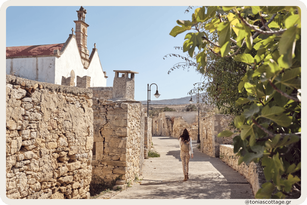 A woman walking down a narrow alley in Crete, with a church steeple visible in the background