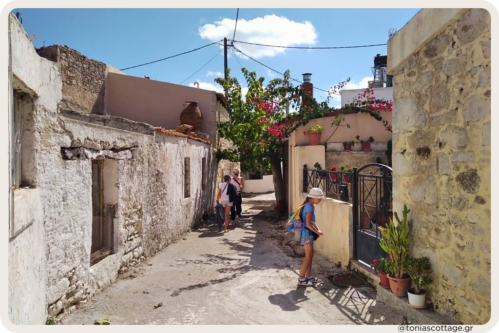 Tourists walking a narrow bougainvillea-lined alley in a sunlit Cretan village