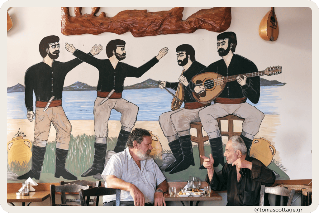 Two men sitting in a kafenio, talking, drinking coffee, with Cretan dancers painted on the wall