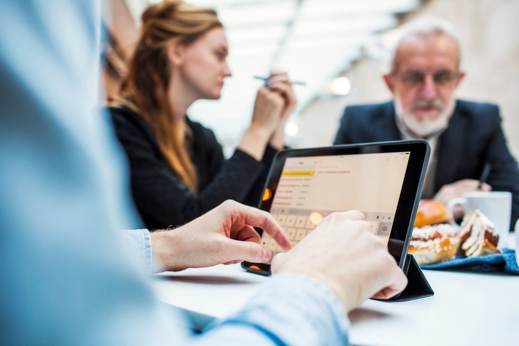 Person using a tablet during a meeting, with colleagues in discussion in the background.