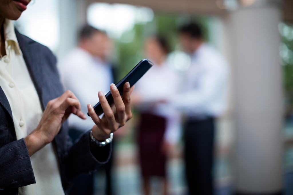 Person using a smartphone with colleagues conversing in the background.