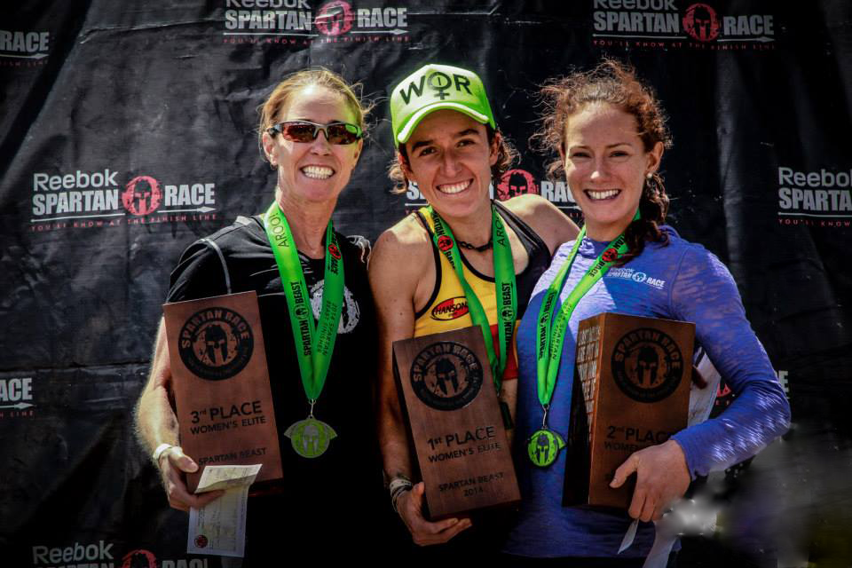 Three female athletes holding trophies on the Reebok Spartan Race podium after finishing the Beast Women's Elite.