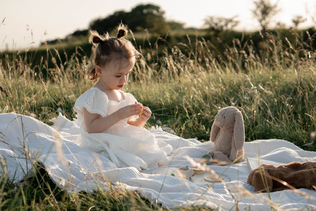 Toddler girl in white dress having a picnic with a stuffed bunny in a sunny grassy field.