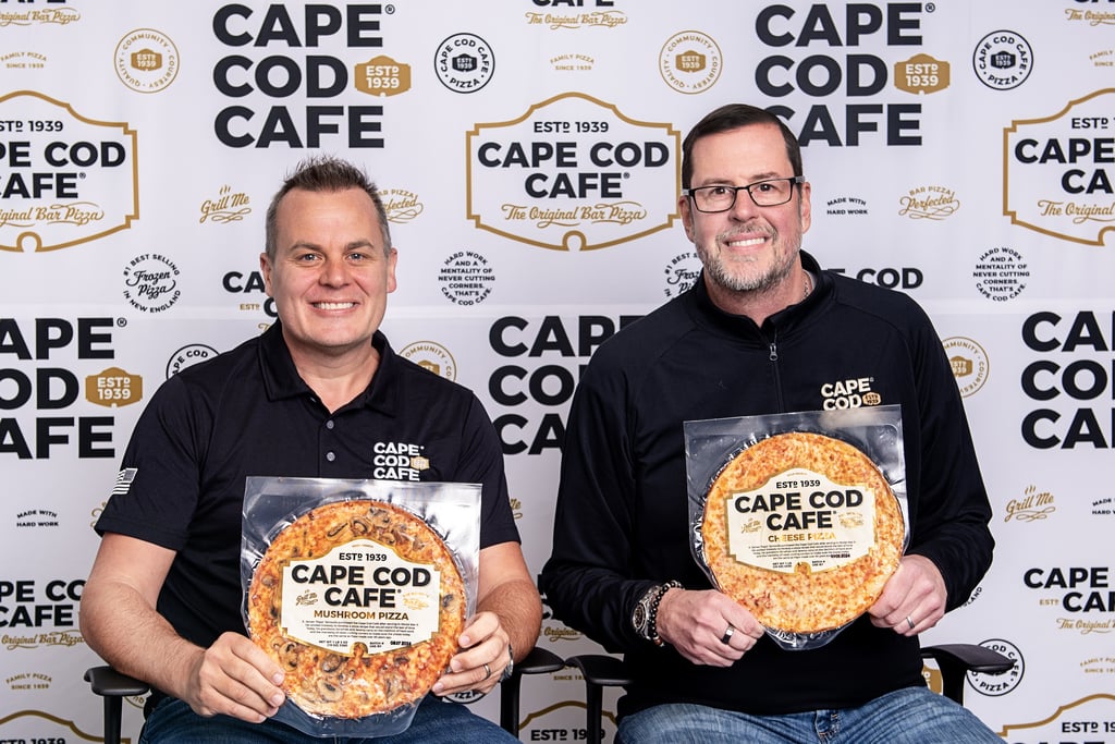 Two men hold Cape Cod Cafe frozen bar pizzas in front of a company logo step and repeat backdrop.