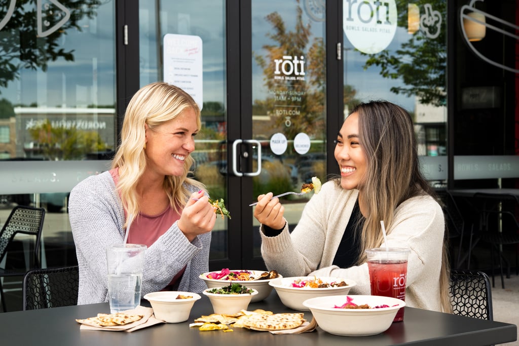 Two women smiling and enjoying Mediterranean bowls and pita bread at an outdoor Roti restaurant table.