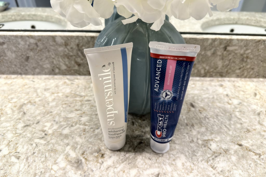 Two tubes of toothpaste on a clean bathroom counter beside a vase of flowers in soft natural light