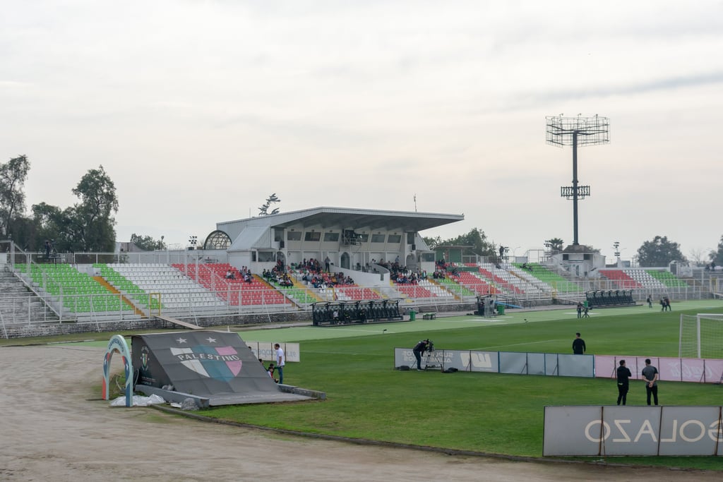 Vista lateral da arquibancada do Estádio Municipal de La Cisterna sob céu azul, com gramado e redes de proteção.