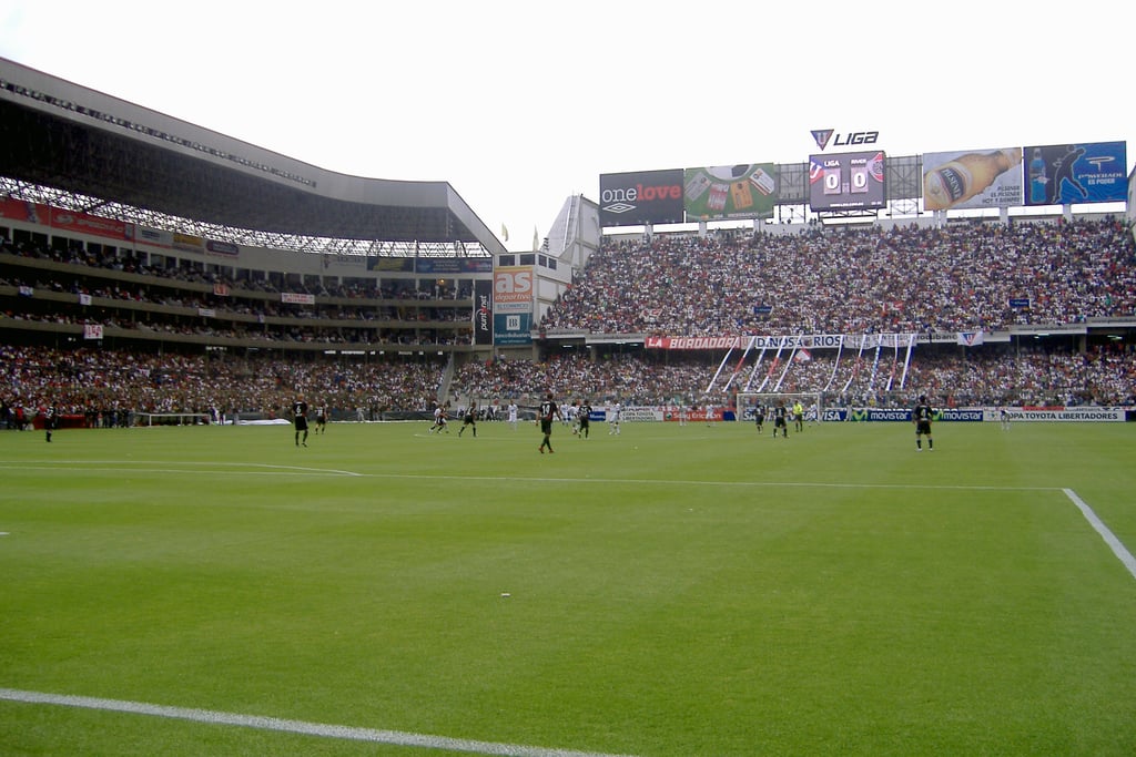 Vista interna do Estádio Rodrigo Paz Delgado em Quito, com arquibancadas brancas e gramado durante uma partida.
