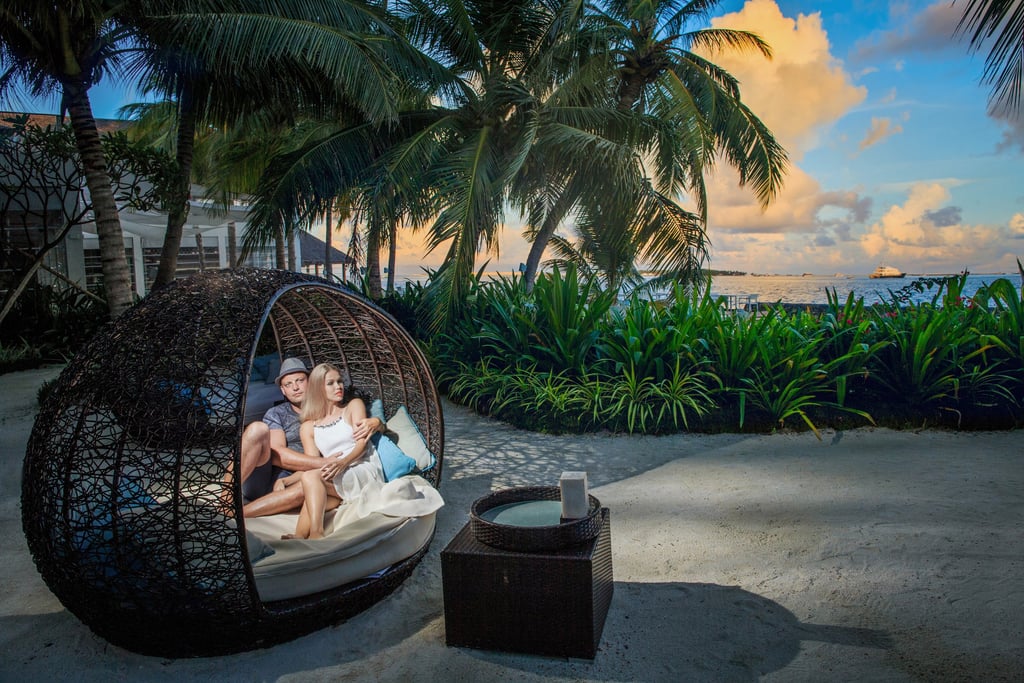 A romantic couple sitting in a cozy, woven wicker pod under tropical palm trees, enjoying