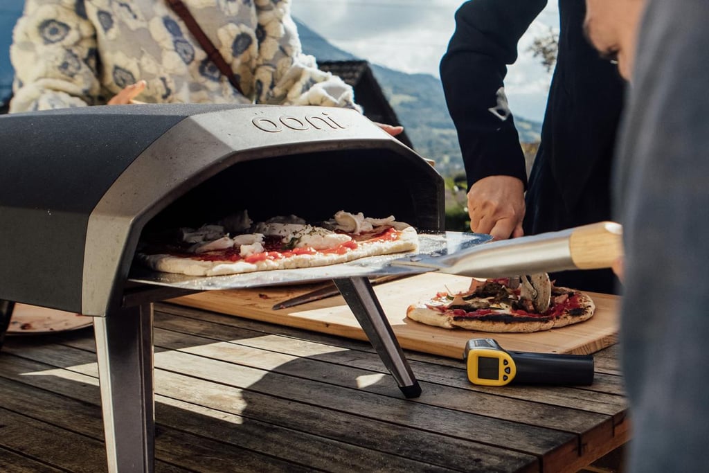 Fresh homemade pizza being slid into a portable Ooni outdoor pizza oven with a metal peel.