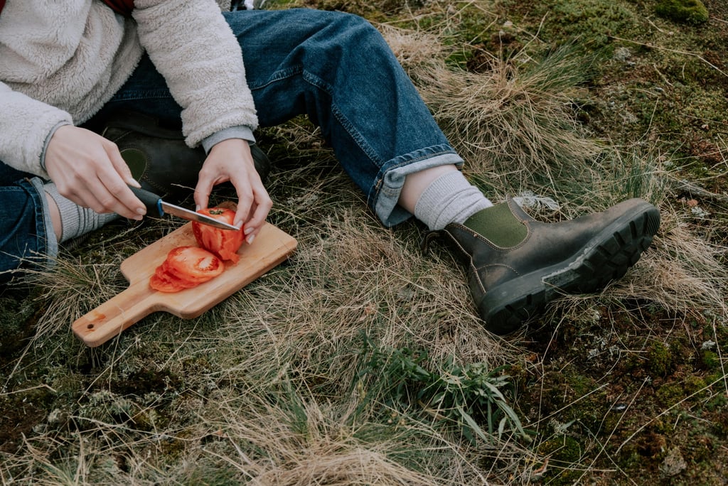 A pereson sitting on the grass cutting a tomato on a cutting board.