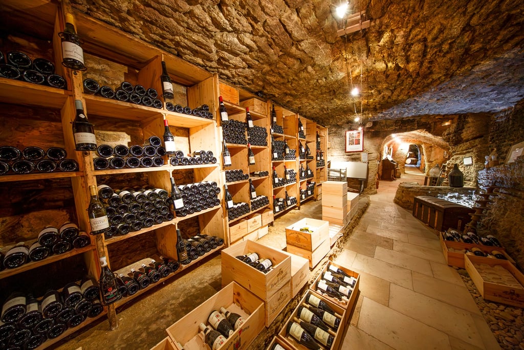 Rustic underground wine cellar with stone walls and wooden shelves storing vintage wine bottles.