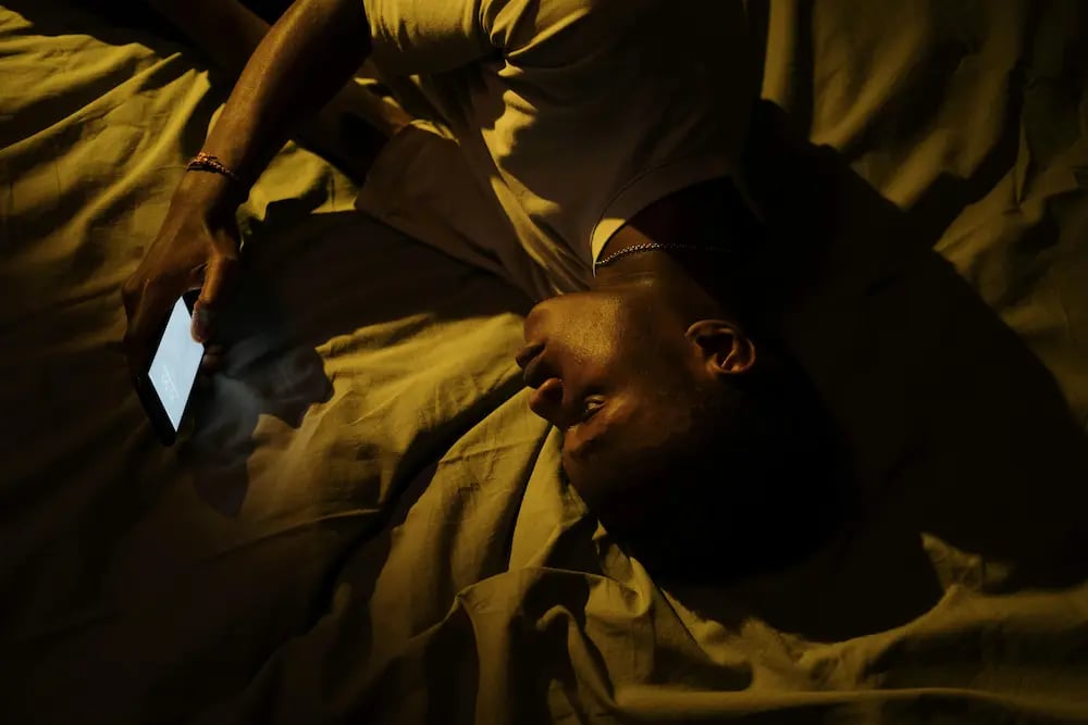A man lying in bed at night using a glowing smartphone with dim, warm lighting.