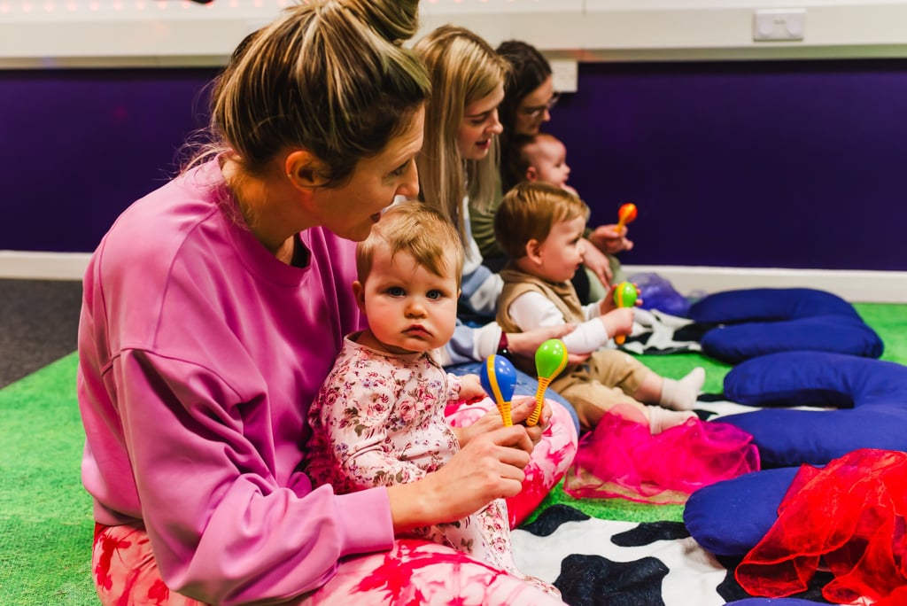 Young girl sat on mums lap playing with shaker instruments. Other children with their mums too.