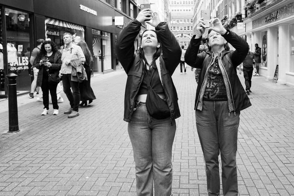 Two female tourists in Carnaby Street London look up towards the sky and simultaneously take photos