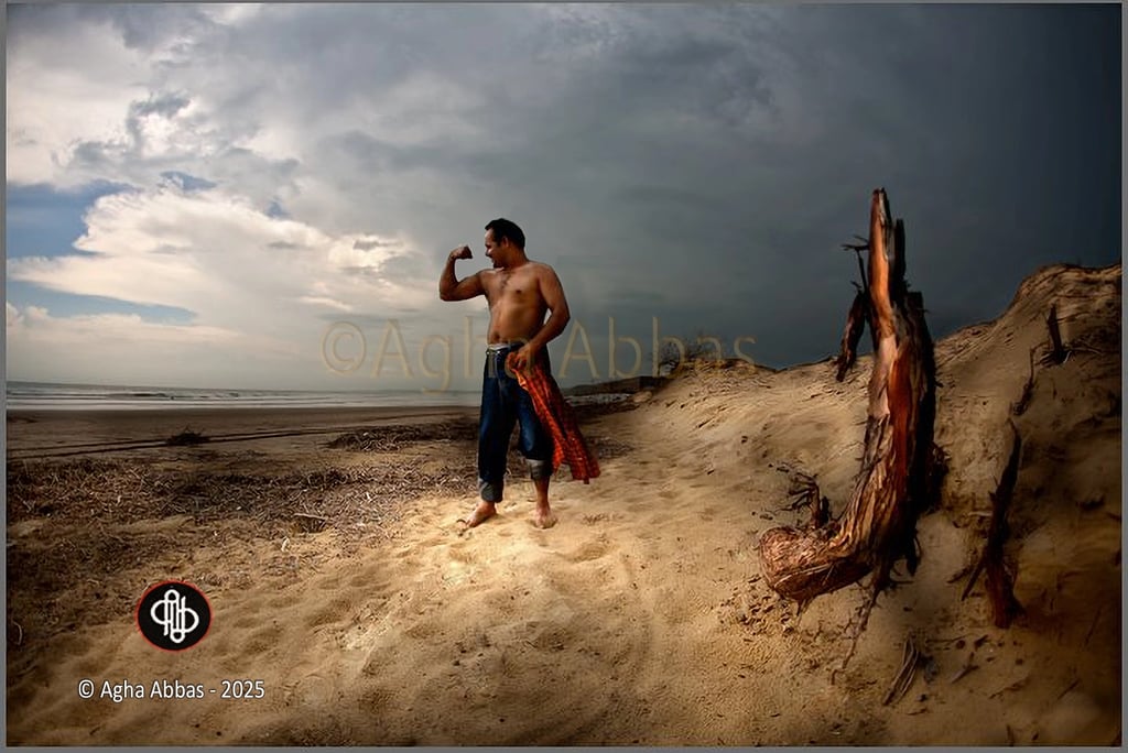 Shirtless barefoot man flexing on beach with jeans and plaid shirt, driftwood, ocean, cloudy sky.