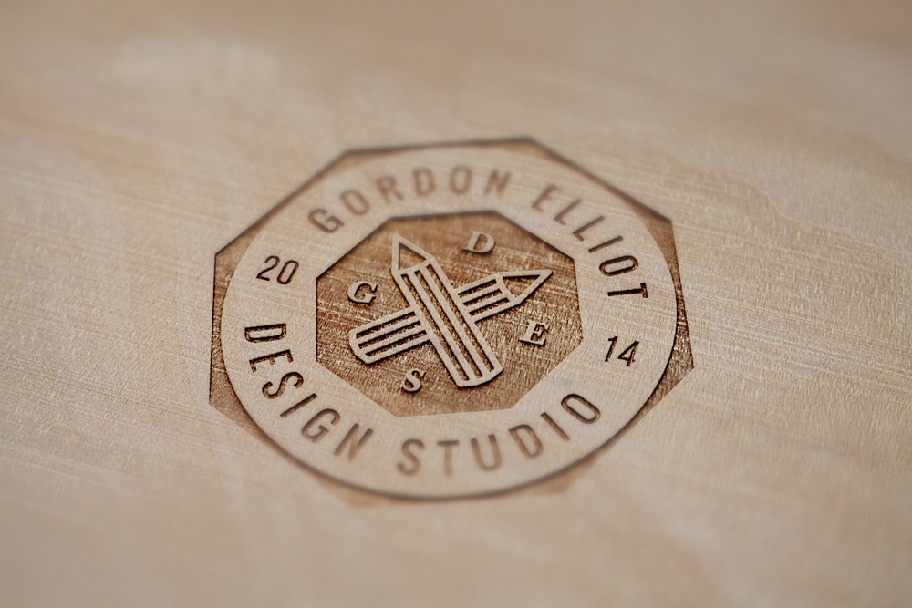 Laser engraved Gordon Elliot Design Studio logo on a light wood surface featuring crossed pencils.