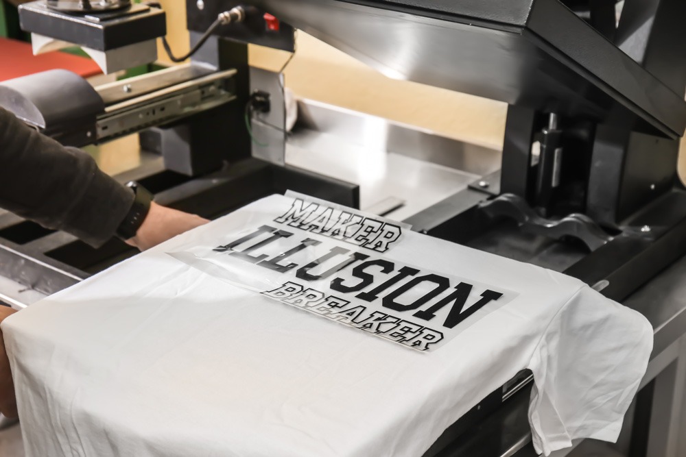 A heat press machine applying a custom vinyl decal to a white t-shirt in a printing shop.