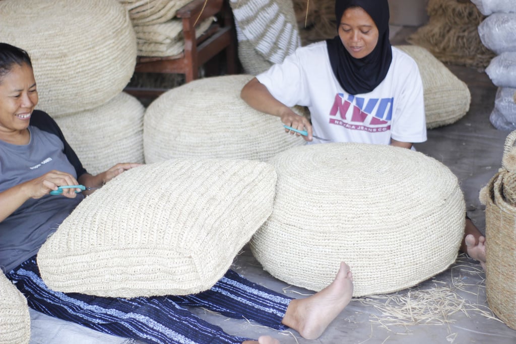 Two Indonesian artisans weaving and trimming handmade straw floor poufs and seagrass cushions.
