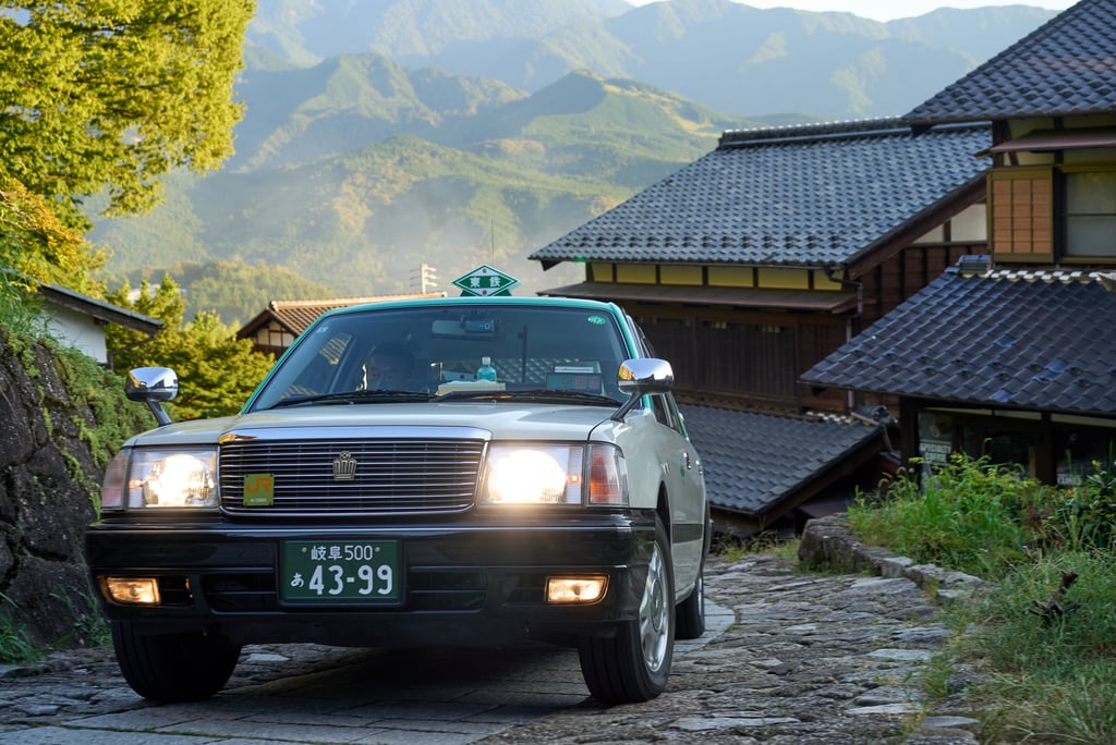 A vintage Japanese Toyota Crown taxi driving on a cobblestone road in a rural mountain village.