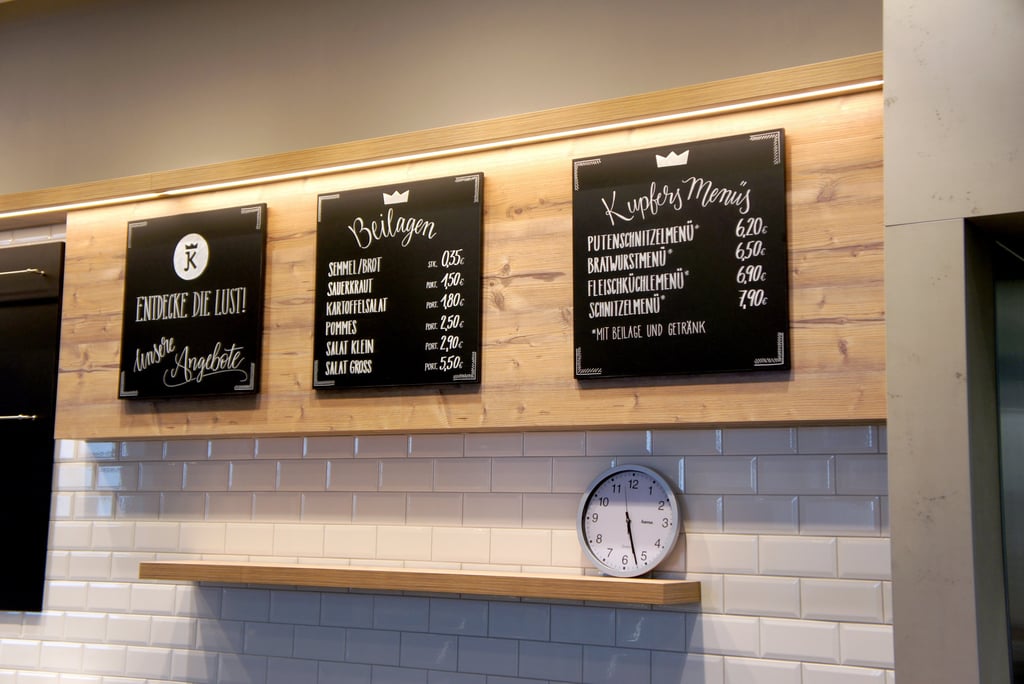 German butcher shop menu boards displaying side dishes and lunch menu prices on a wooden wall above white tiles.