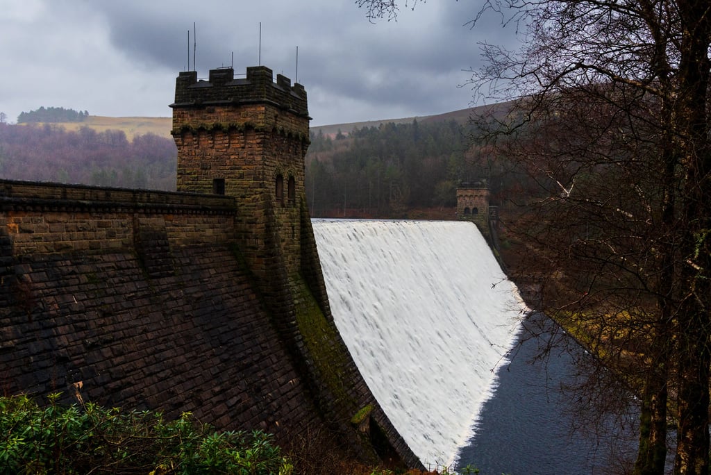 Historic stone Derwent Dam with cascading water overflow and towers in Peak District National Park.