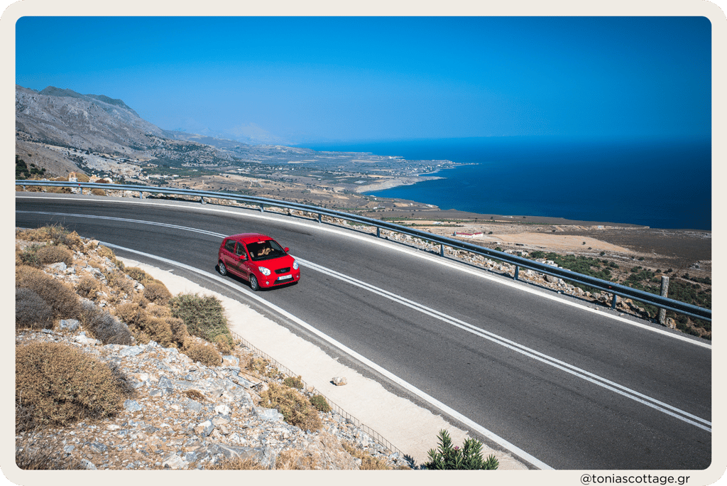 A red rental car driving down a winding road in Crete, Greece, surrounded by beautiful scenery