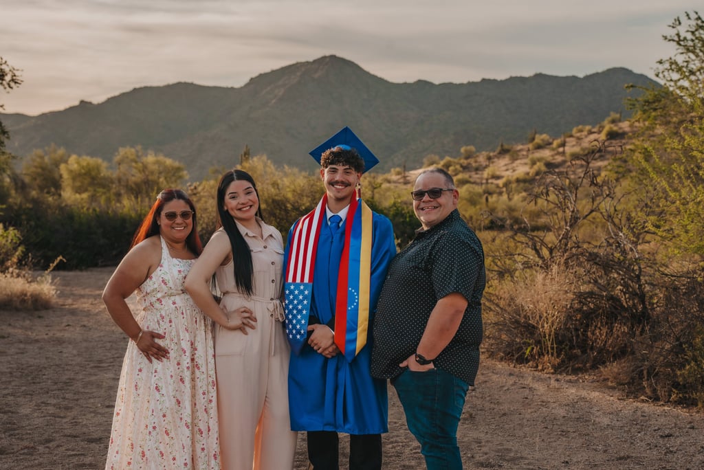A graduate in a blue cap and gown stands with family in a desert landscape at sunset.