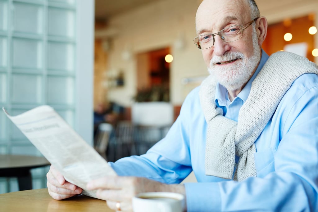 Senhor idoso, de barba, sorridente, de camisa social azul e blusão nas costas lê jornal.