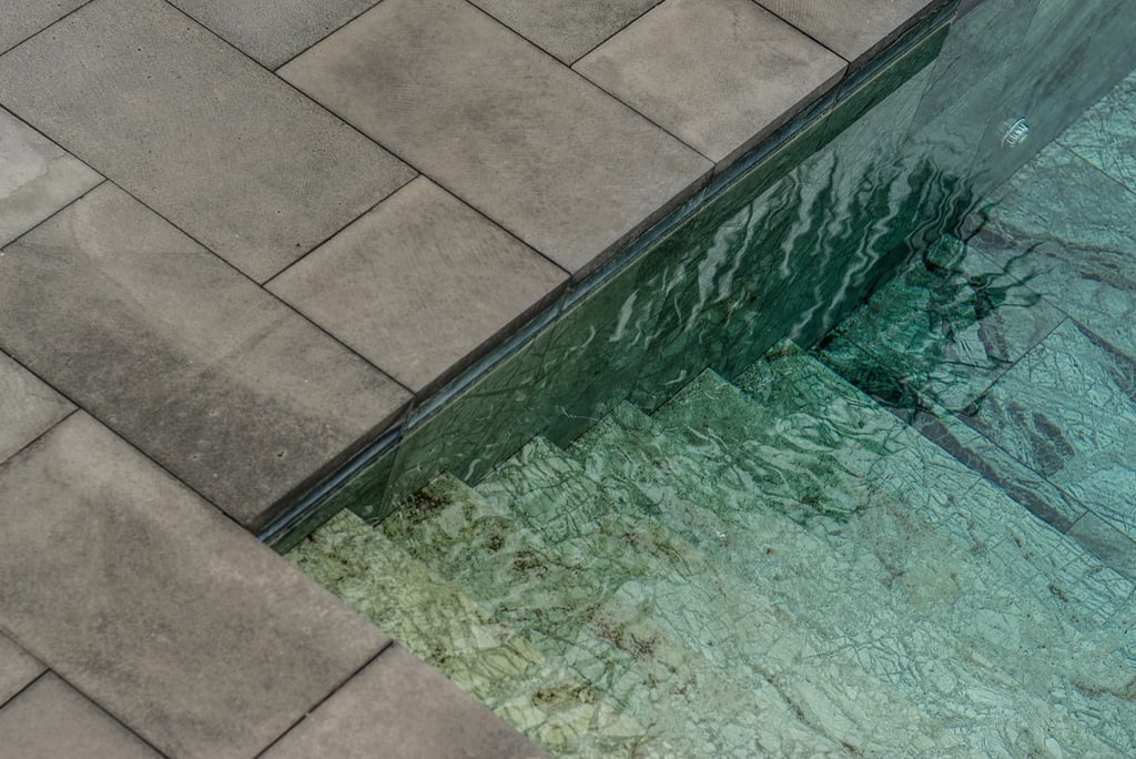 Modern luxury swimming pool with green natural stone tiles and gray concrete paver pool coping.