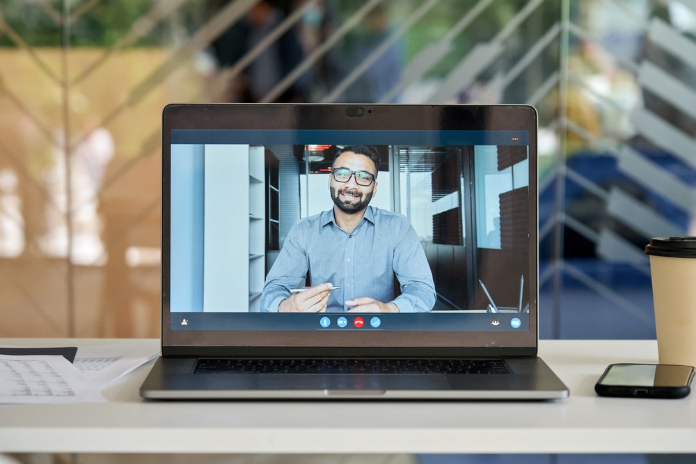A laptop screen showing a video call with a man in a business shirt holding a pen