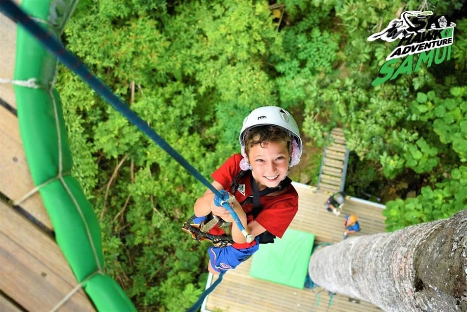 A person is zip-lining across a lush, green landscape, surrounded by dense foliage and rocky outcrop