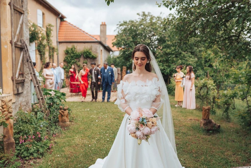 La mariée avance les yeux baissés pendant que ses proches la suivent dans un jardin verdoyant.