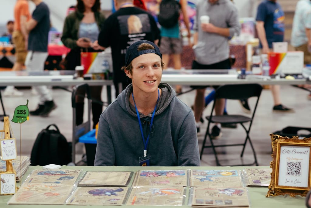 A smiling vendor sits at a table in front of his trading cards for sale.
