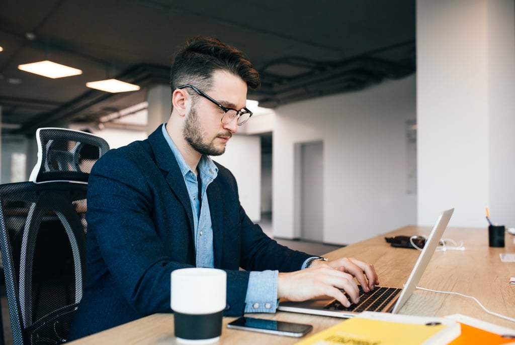 a man in a navy jacket and chambray shirt, sitting at a desk, working on a laptop computer