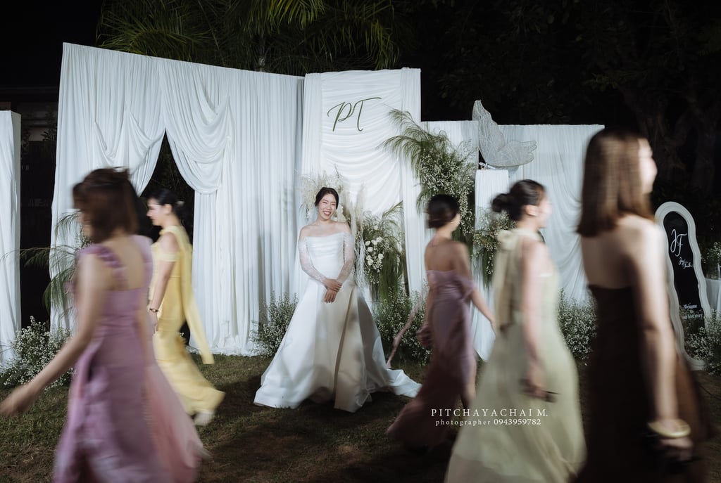 เจ้าสาวยืนกลางฉากงานแต่งโทนขาว ขณะเพื่อนเจ้าสาวเดินผ่านสร้างเอฟเฟกต์เบลอ ภาพงานแต่ง Outdoor ตอนกลางคืน