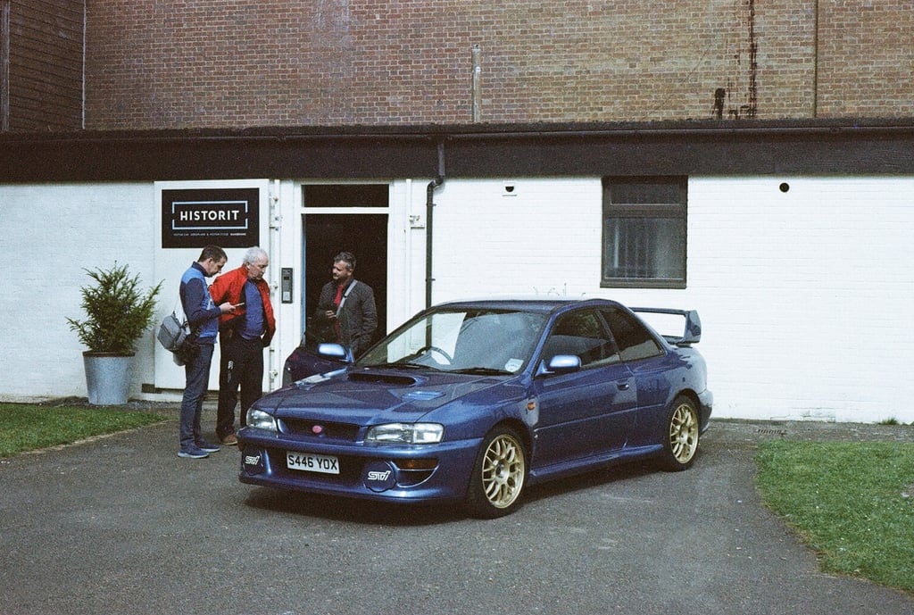 a classic Subaru Impreza parked in front a building with three guys looking at it