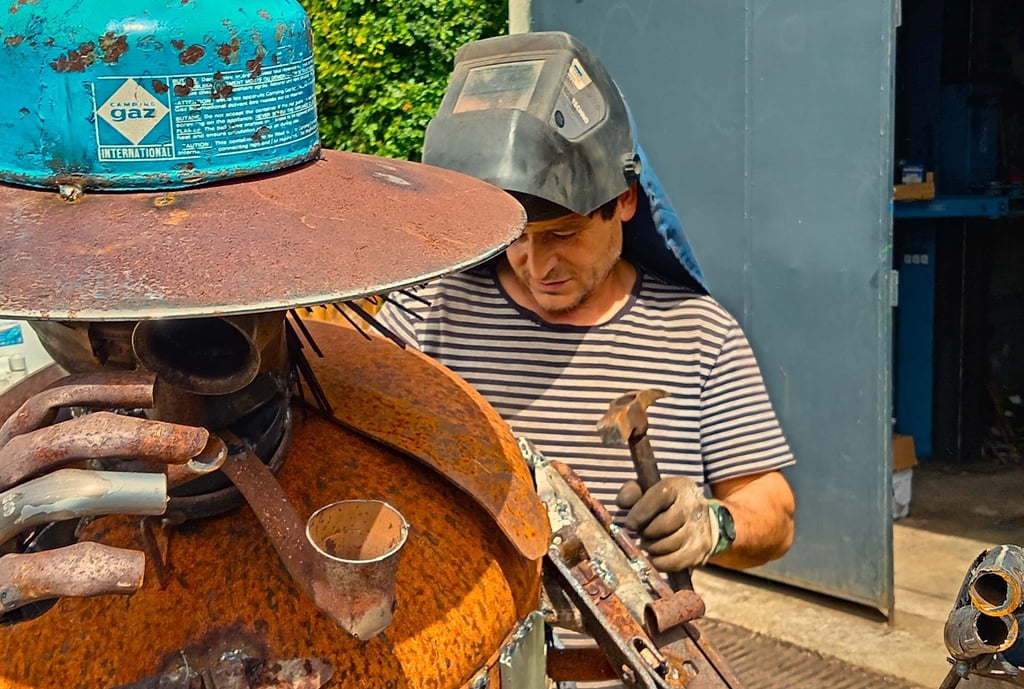 Murat wearing a welding mask and gloves uses a hammer to shape a rusty scrap metal sculpture.